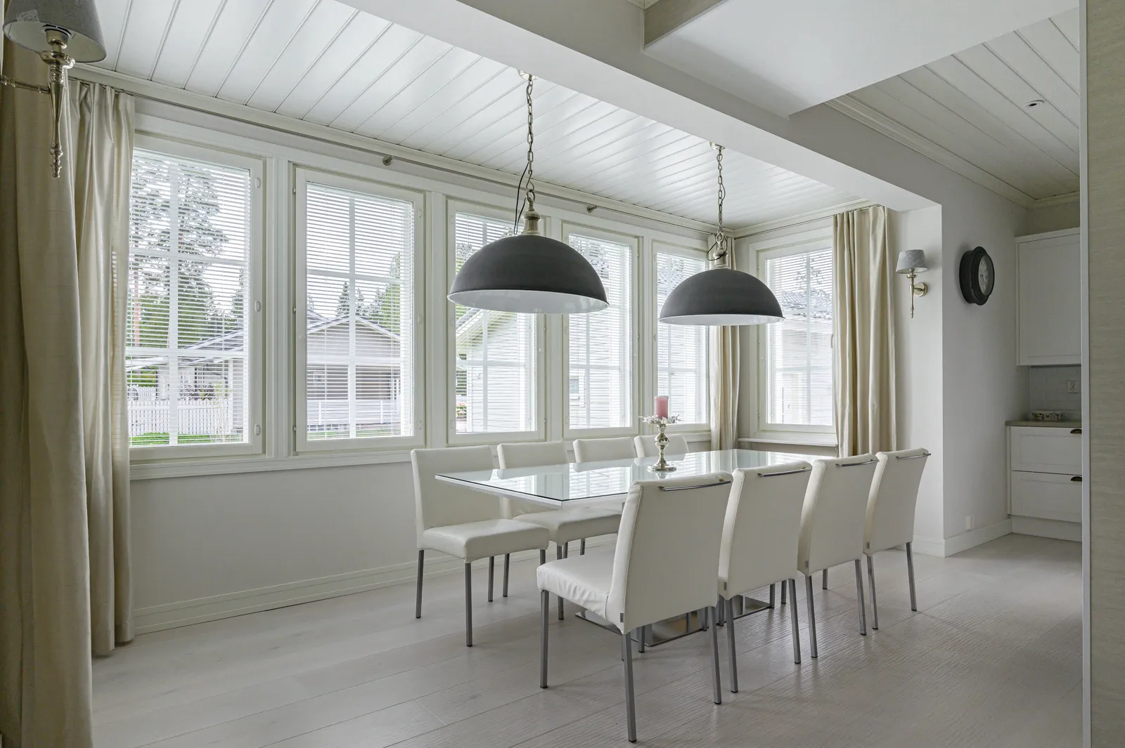 Vaaleassa ruokasalissa on lasipöytä, valkoiset tuolit, suuret ikkunat ja kaksi riippuvalaisinta.