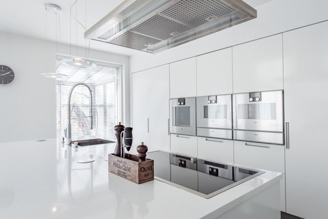 Moderni, valkoinen keittiö, jossa on rosteriset uunit, liesi ja puinen mausteteline kiiltävällä saarekkeella.