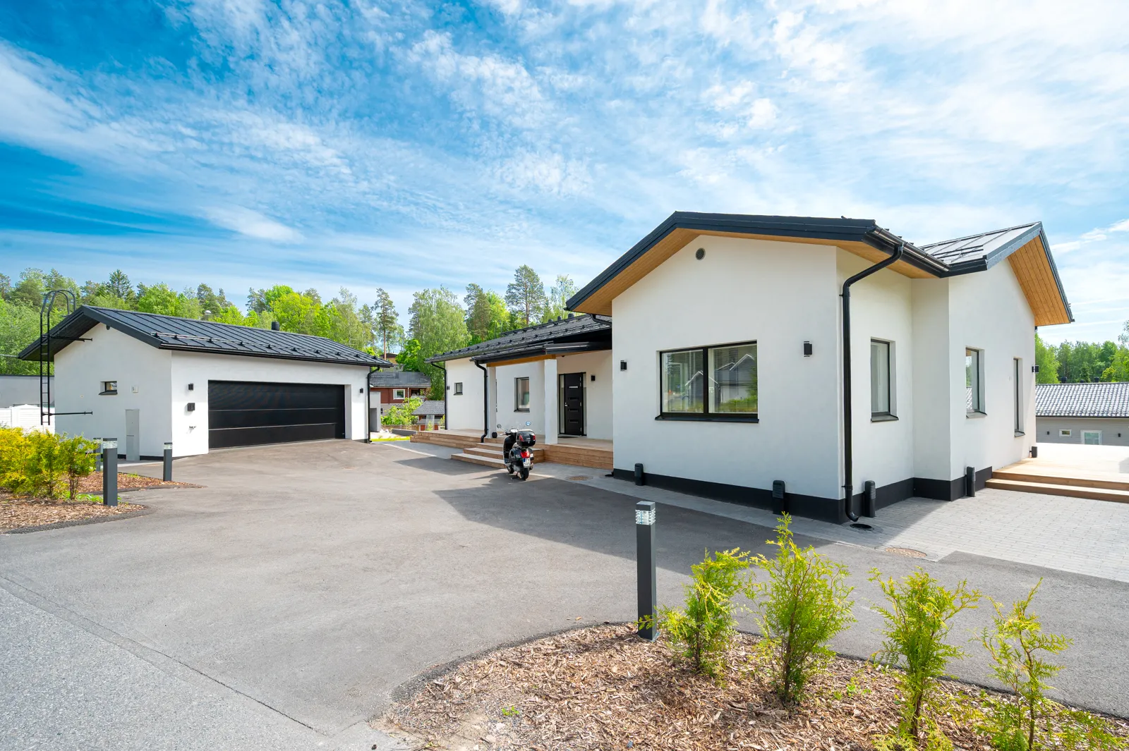 Moderni, valkoinen omakotitalo, jossa on musta katto ja autotalli, aurinkoisena päivänä. Etualalla on asfaltoitu piha, jossa on pieniä vihreitä pensaita ja puuainesta. Taustalla näkyy vihreää metsää ja sininen taivas pilvillä.