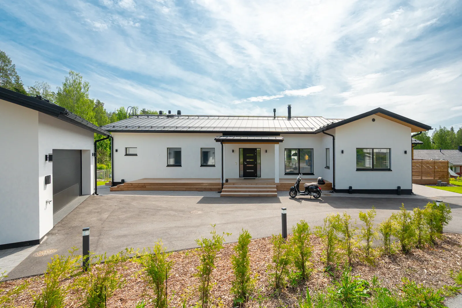 Moderni valkoinen omakotitalo, jossa on tumma katto ja musta skootteri edessä aurinkoisena päivänä.