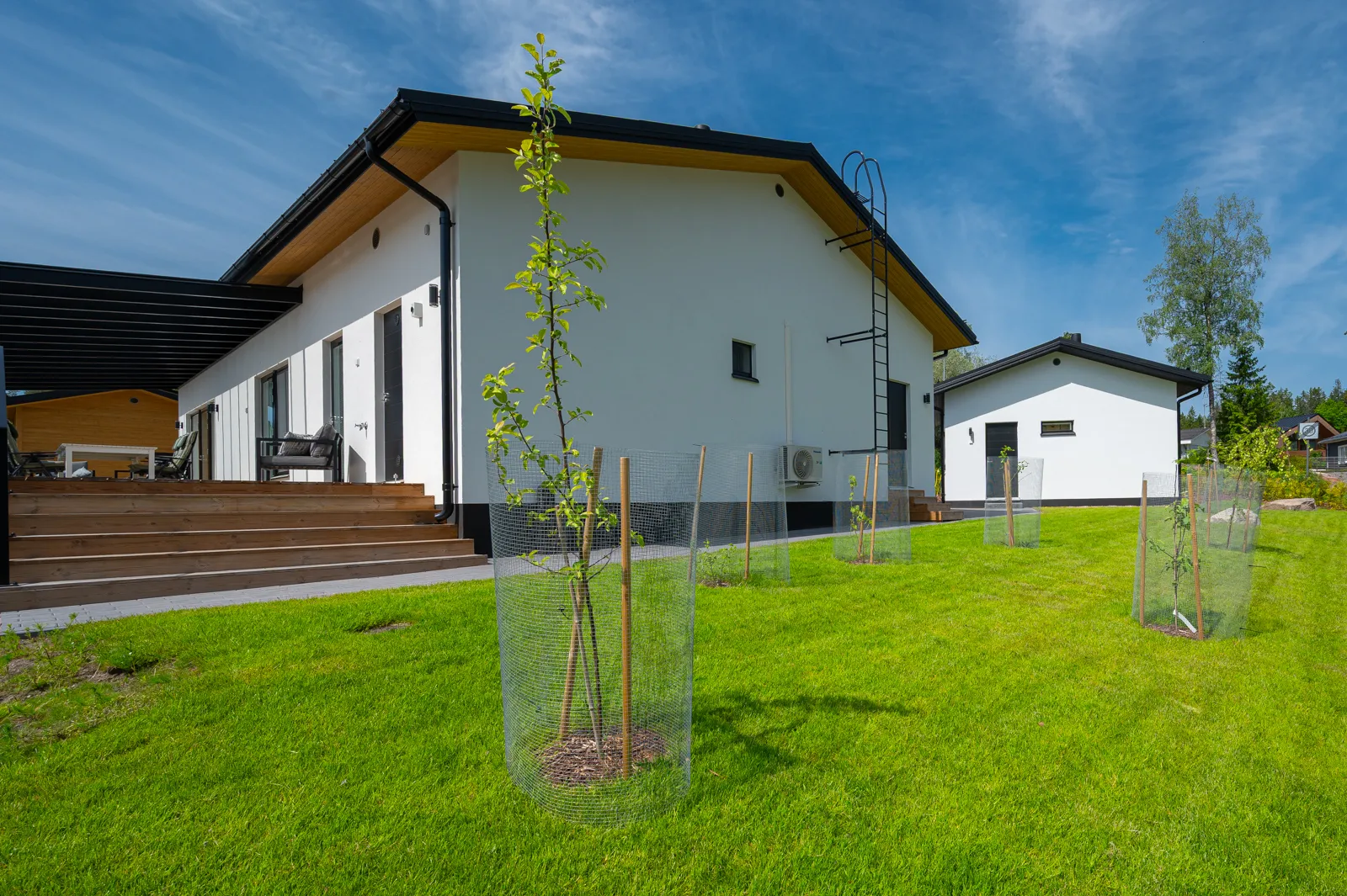 Moderni, valkoinen talo, jossa on musta katto ja puuaita ympärillä, aurinkoisena päivänä.