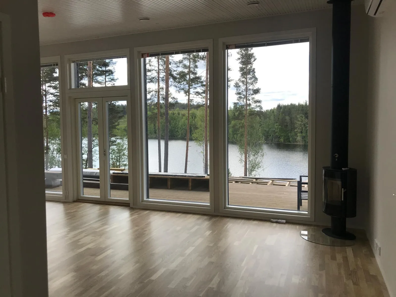 Valoisa huone, jossa on suuret ikkunat, joista näkyy järvi ja metsä, sekä musta takka.