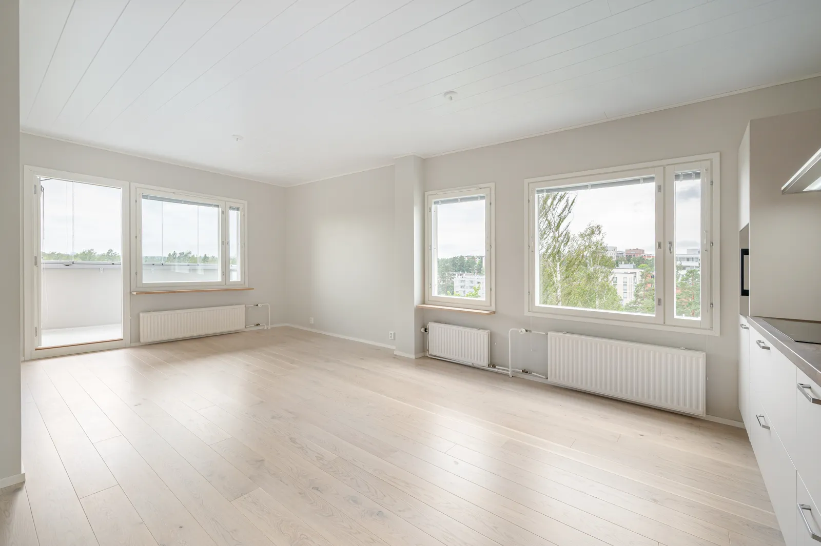 Vaalea ja avara huoneisto, jossa on suuret ikkunat, valkoiset seinät ja vaalea puulattia. Huoneessa on patterit ikkunoiden alla ja parveke.