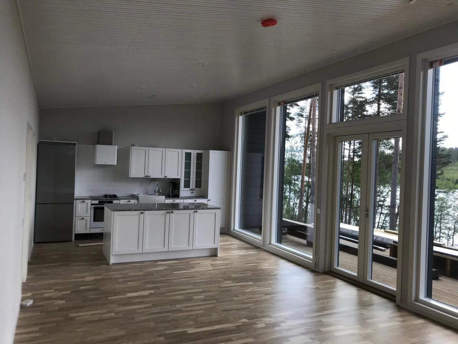 Vaaleassa huoneessa on valkoinen keittiö, suuret ikkunat ja puulattia, luoden avaran ja valoisan tilan.