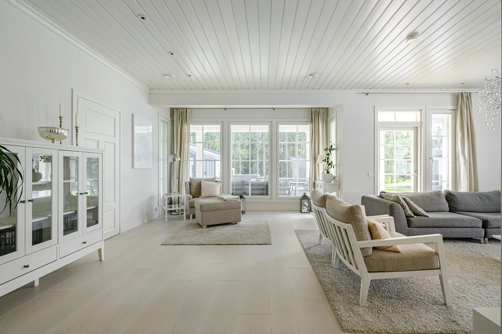 Vaaleassa olohuoneessa on valkoinen katto, suuret ikkunat, vaaleat huonekalut ja harmaa sohva. Huoneessa on myös valkoinen kaappi ja mattoja. Valoisa ja avara tunnelma.