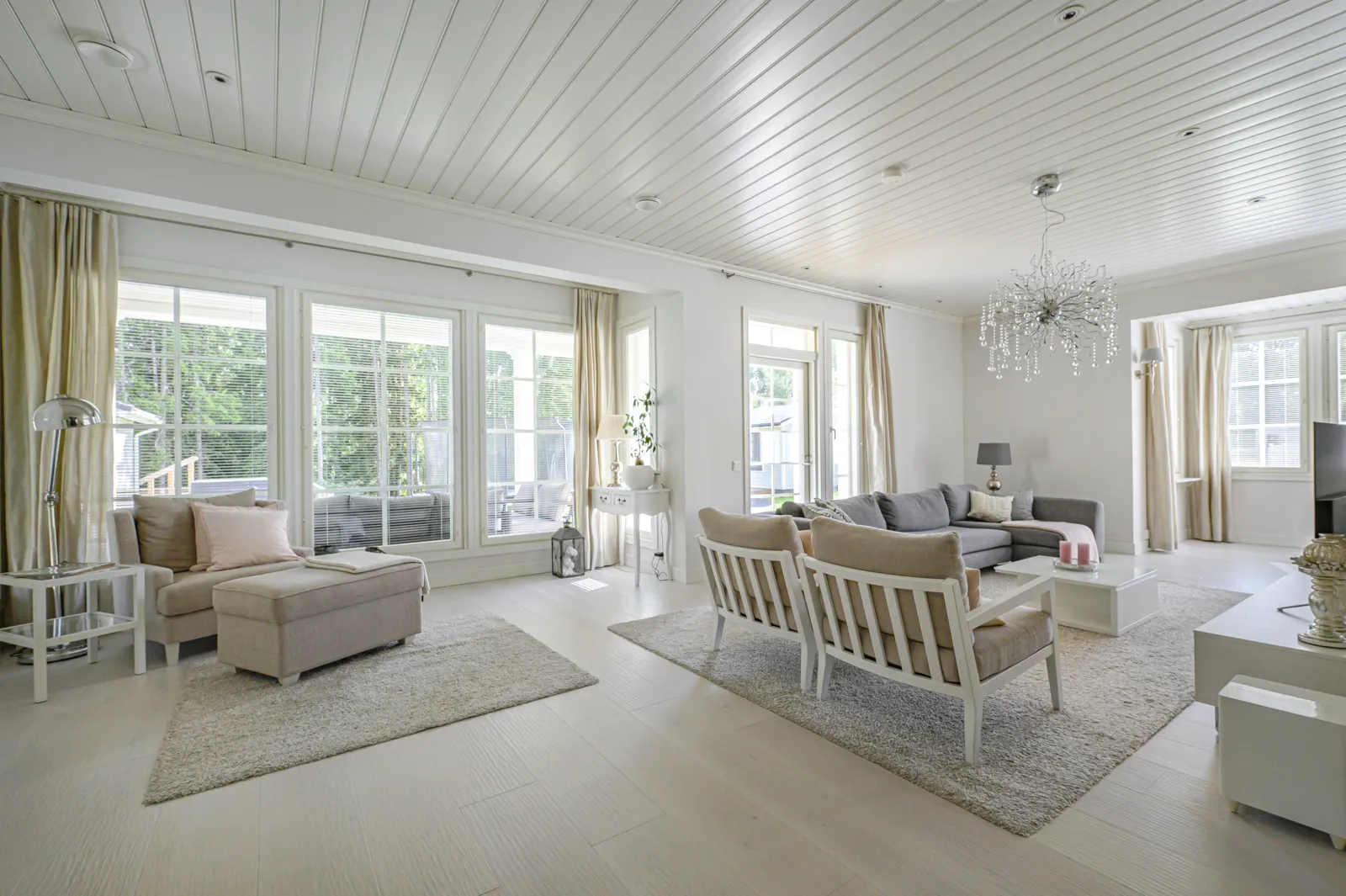 Vaalea olohuone, jossa on suuret ikkunat, harmaa sohva, valkoinen katto ja kristallikruunu.