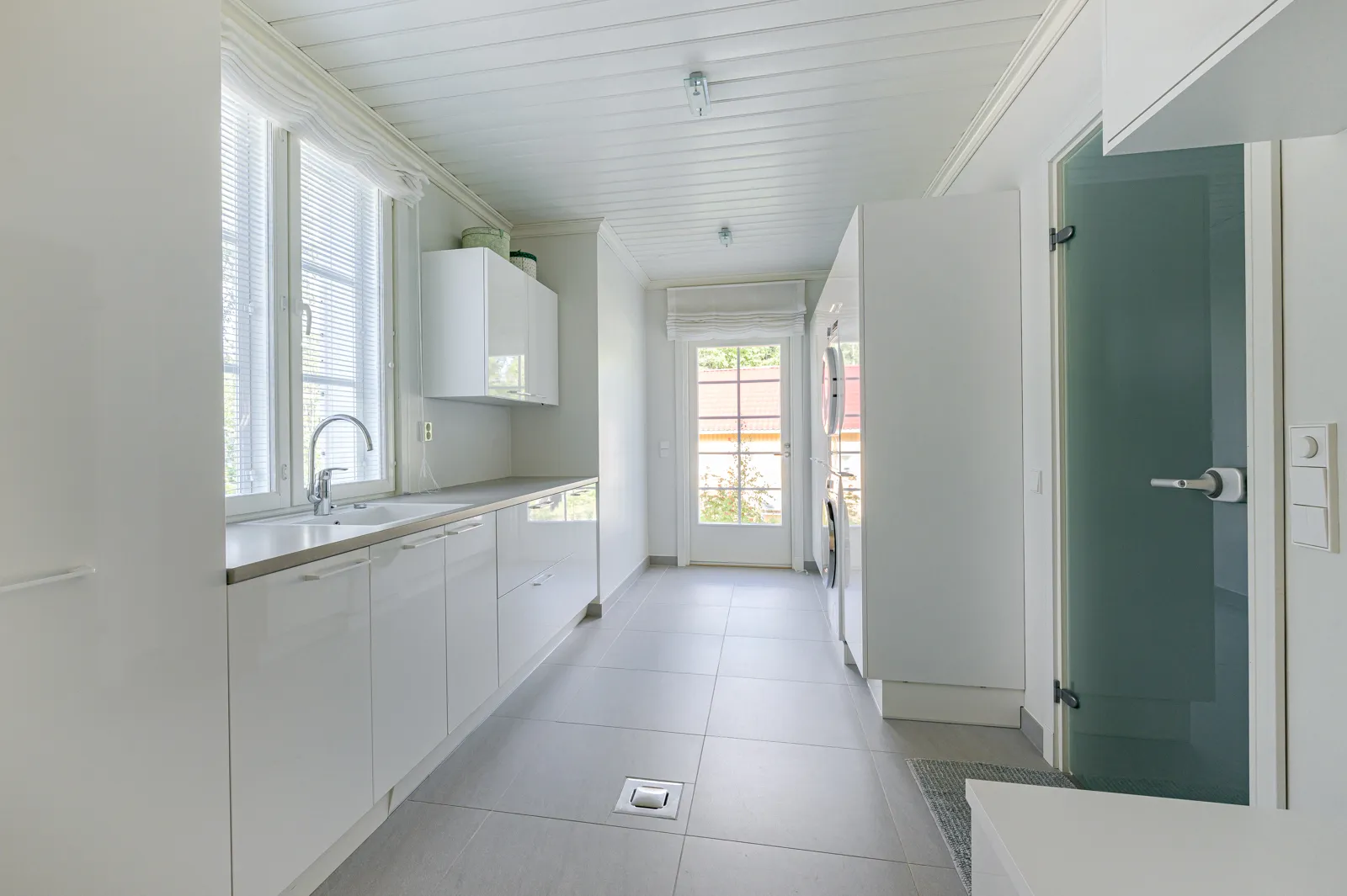 Vaaleassa kodinhoitohuoneessa on valkoiset kaapit, harmaat laatat ja ikkunasta tulviva luonnonvalo.