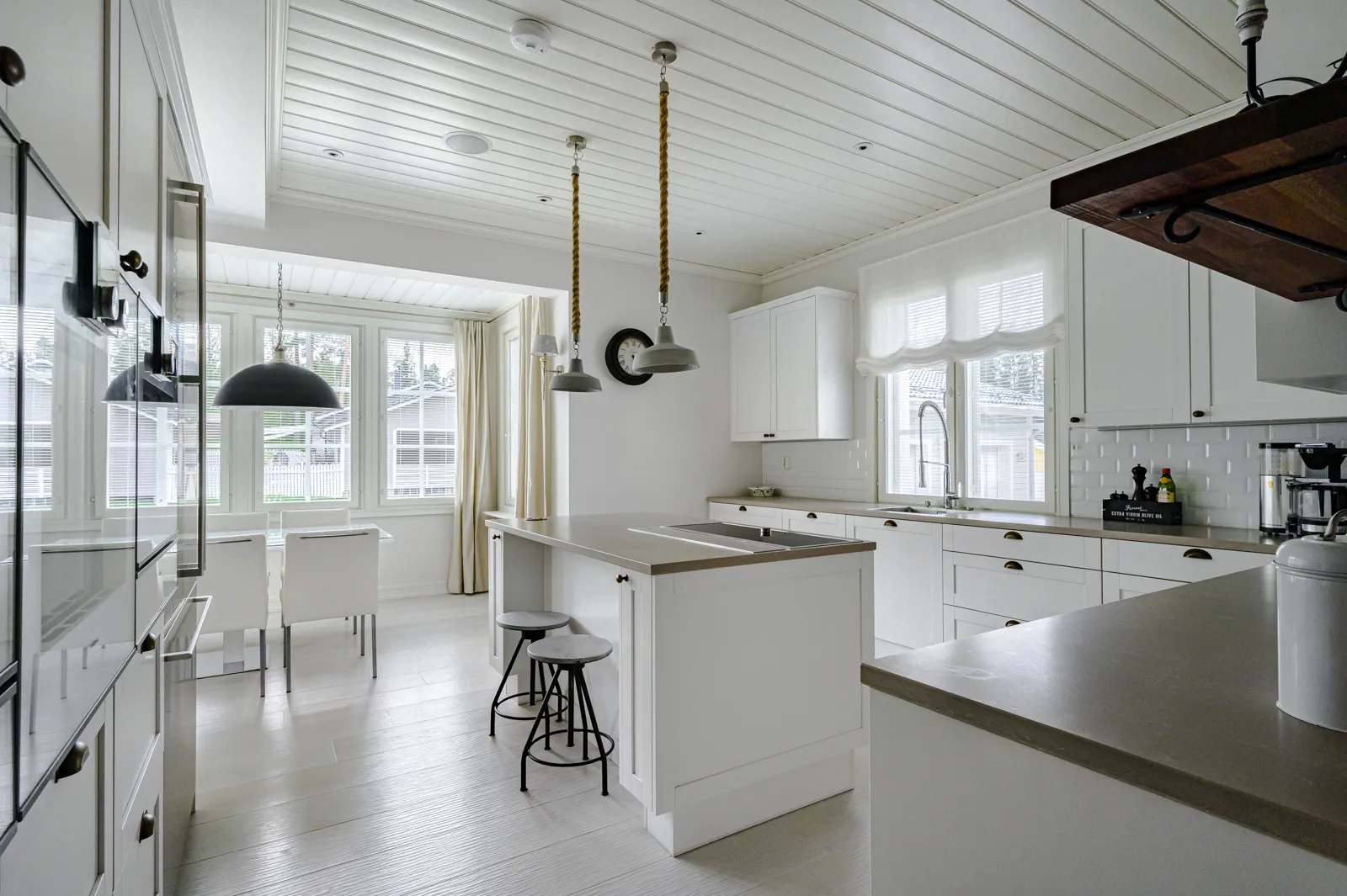 Valkoinen keittiö, jossa on saareke, kaksi roikkuvaa valaisinta ja ikkunat, jotka tuovat luonnonvaloa.