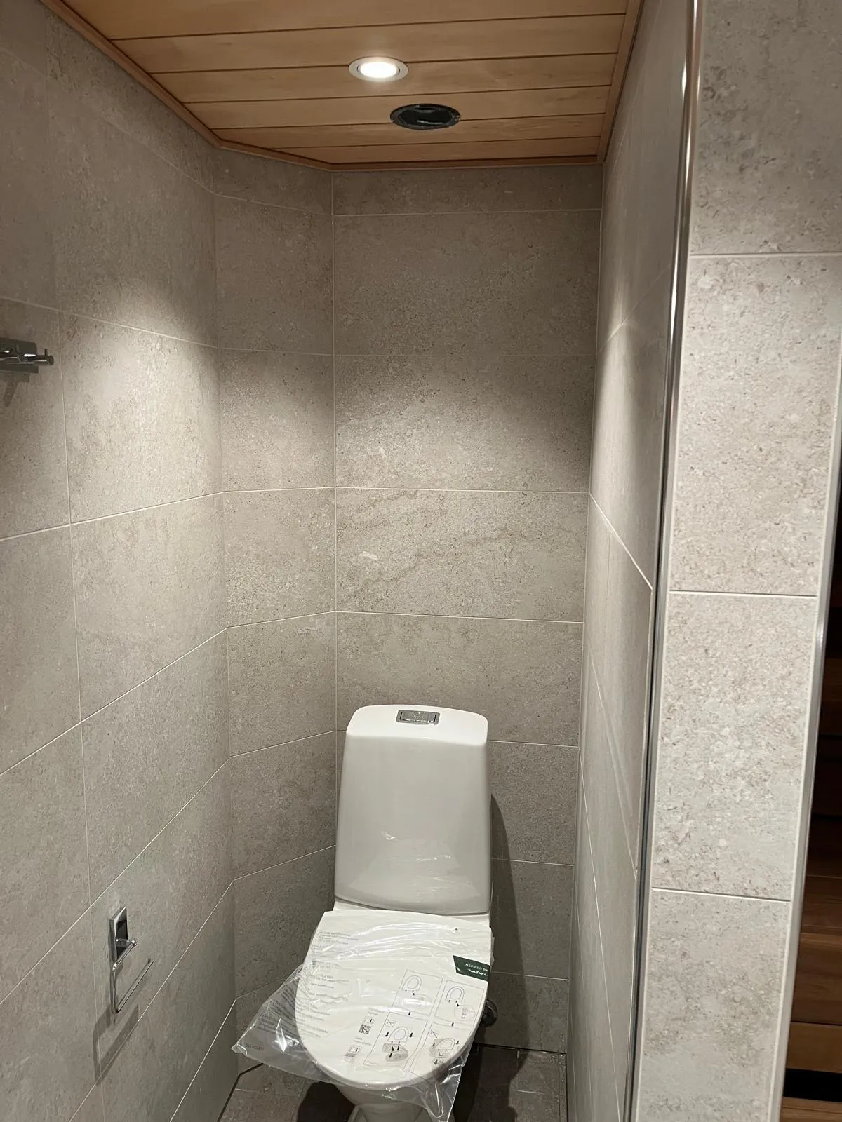 Kylpyhuoneen kuva, jossa on valkoinen wc-istuin harmaiden laattojen ja puisen katon alla.