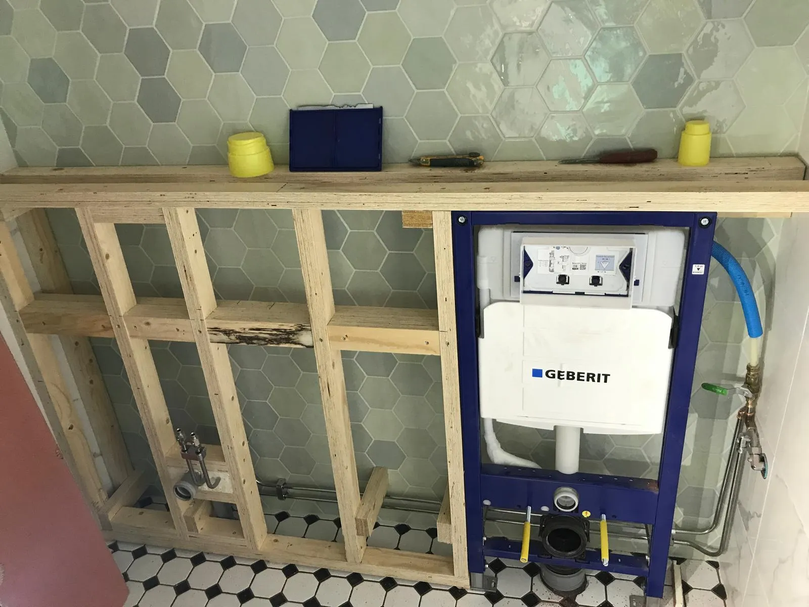 Kylpyhuoneen seinään asennettu puurunko, jonka sisällä on Geberit-merkkinen WC-säiliö, taustalla kuusikulmaisia laattoja ja lattialla mustavalkoisia laattoja.